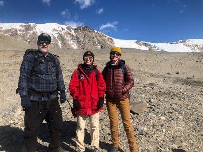 3 people stand on rocky, snowy mountain plateau dressed in cold weather attire